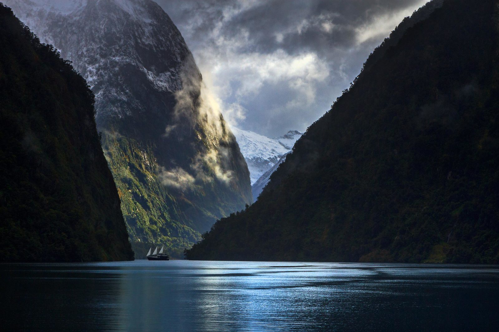 A ship with 3 sails is dwarfed by the immensity of the mountains at Doubtful Sound, New Zealand