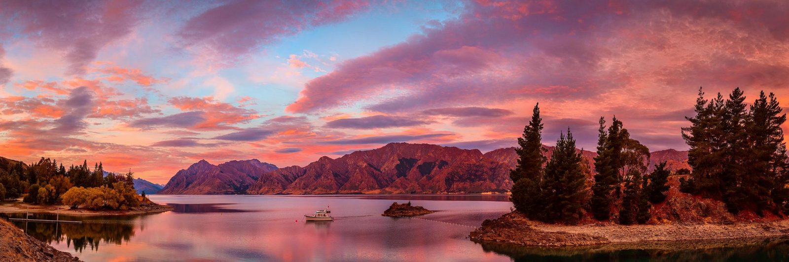 A small boat is dwarfed by the mountains surrounding Lake Hawea, near Wanaka, New Zealand during a very colourful sunrise